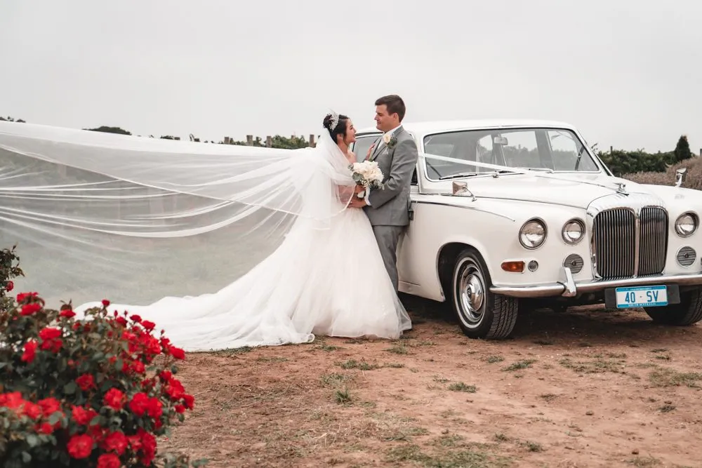 A bride and groom beside a vintage white Daimler, her veil streaming out in the wind, red roses in the foreground