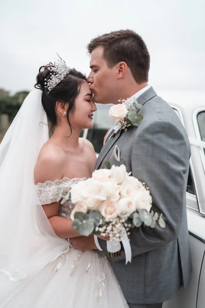 A groom in grey kissing his bride's forehead, she in a tiara with a bouquet of white roses