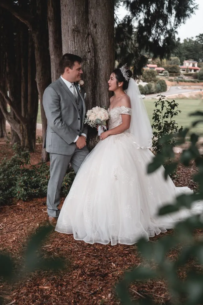 A bride in a full gown and a groom in grey standing hand in hand under tall pines, the vineyard beyond