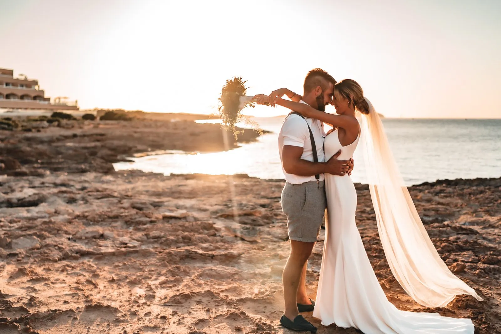 A bride and groom on a rocky shore at sunset, her veil lifting in the wind and the sun flaring behind them