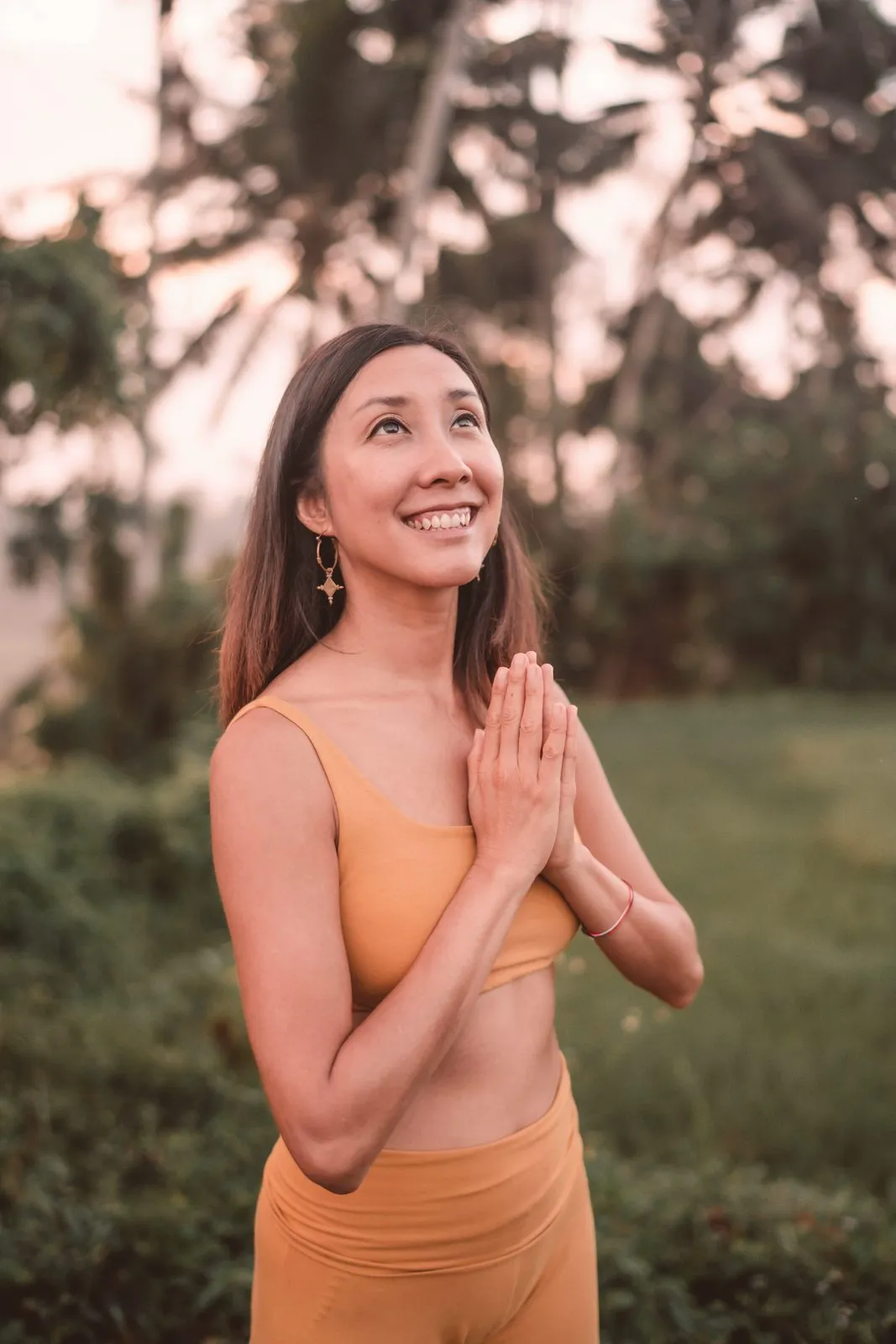 A smiling woman in ochre yoga wear with her hands in prayer, palms and morning mist behind her, Bali