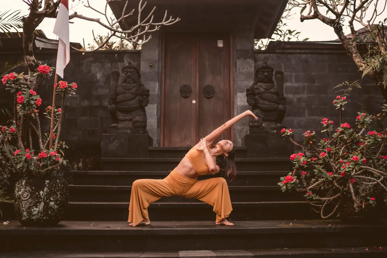 A woman in ochre in a wide-legged forward fold on the steps of a Balinese temple, red hibiscus either side