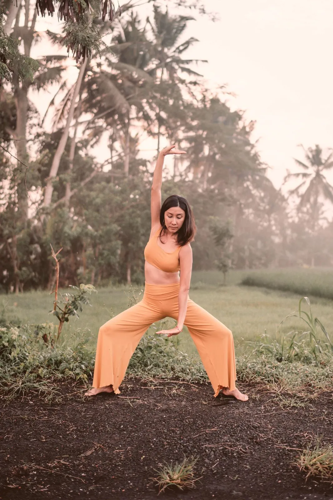 A woman in ochre in goddess pose with one arm reaching up, palm trees and morning haze behind her