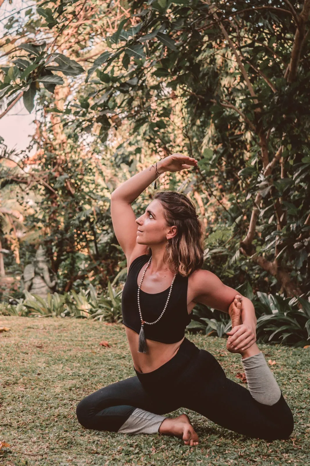 A woman in black in a seated twist on the grass under trees, one arm curved over her head