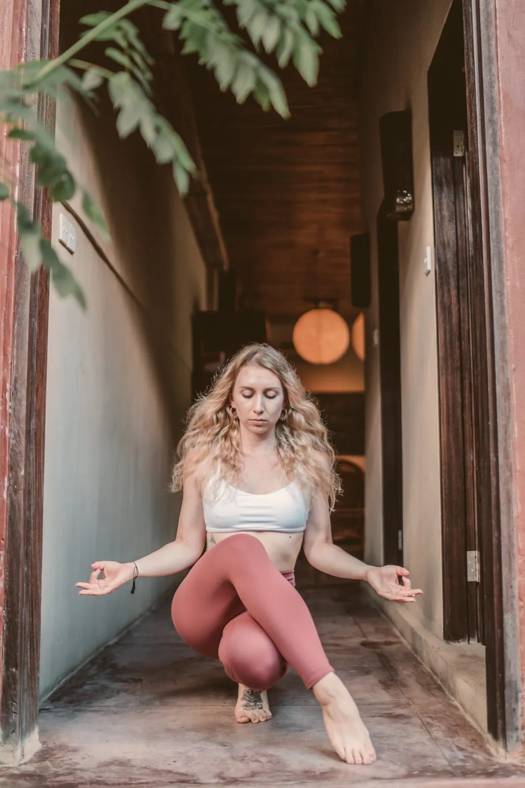 A woman in rust leggings in a low squat in a narrow corridor, hands in mudra, eyes closed