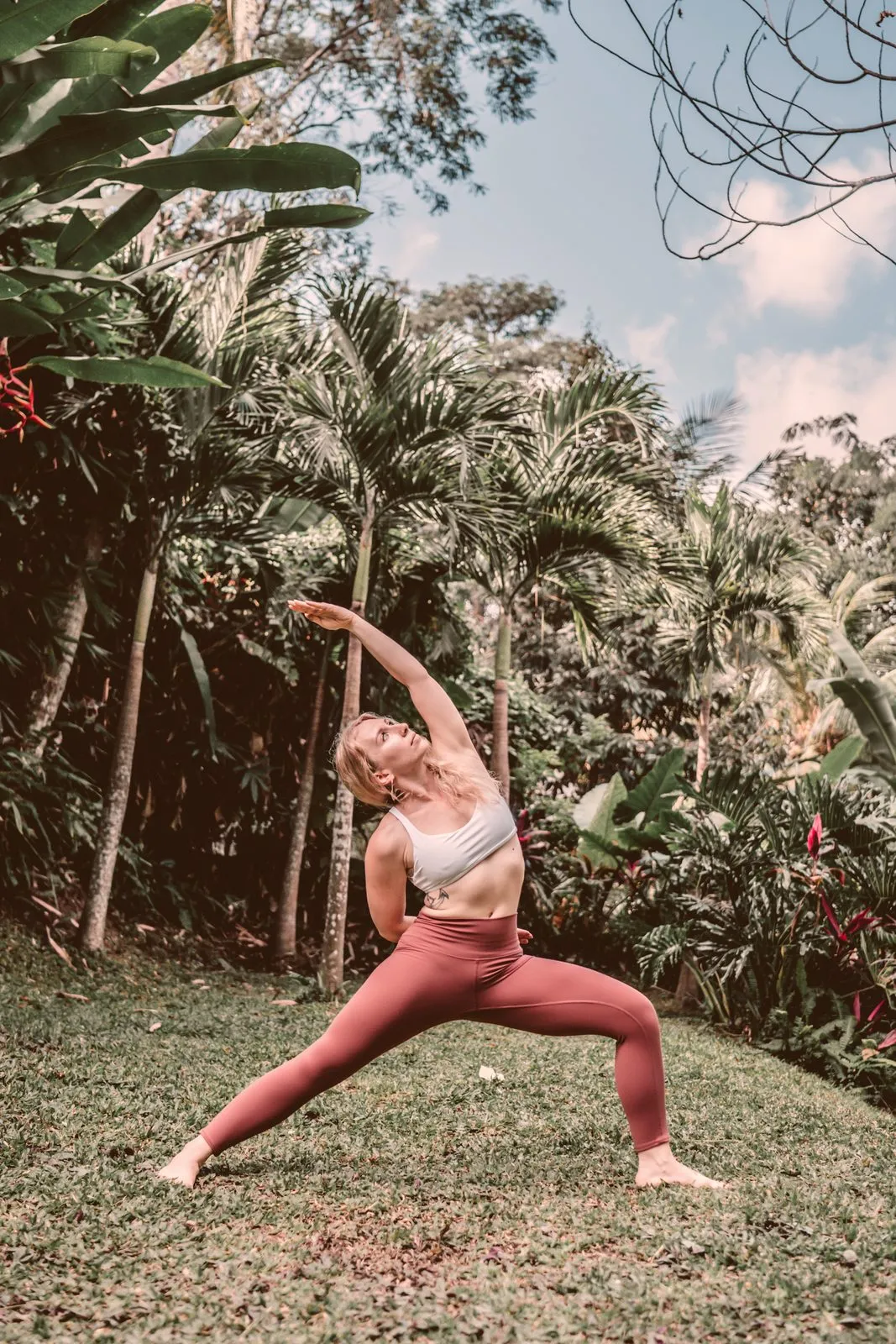 A woman in rust leggings in a high lunge with arms raised on a lawn among palms and frangipani
