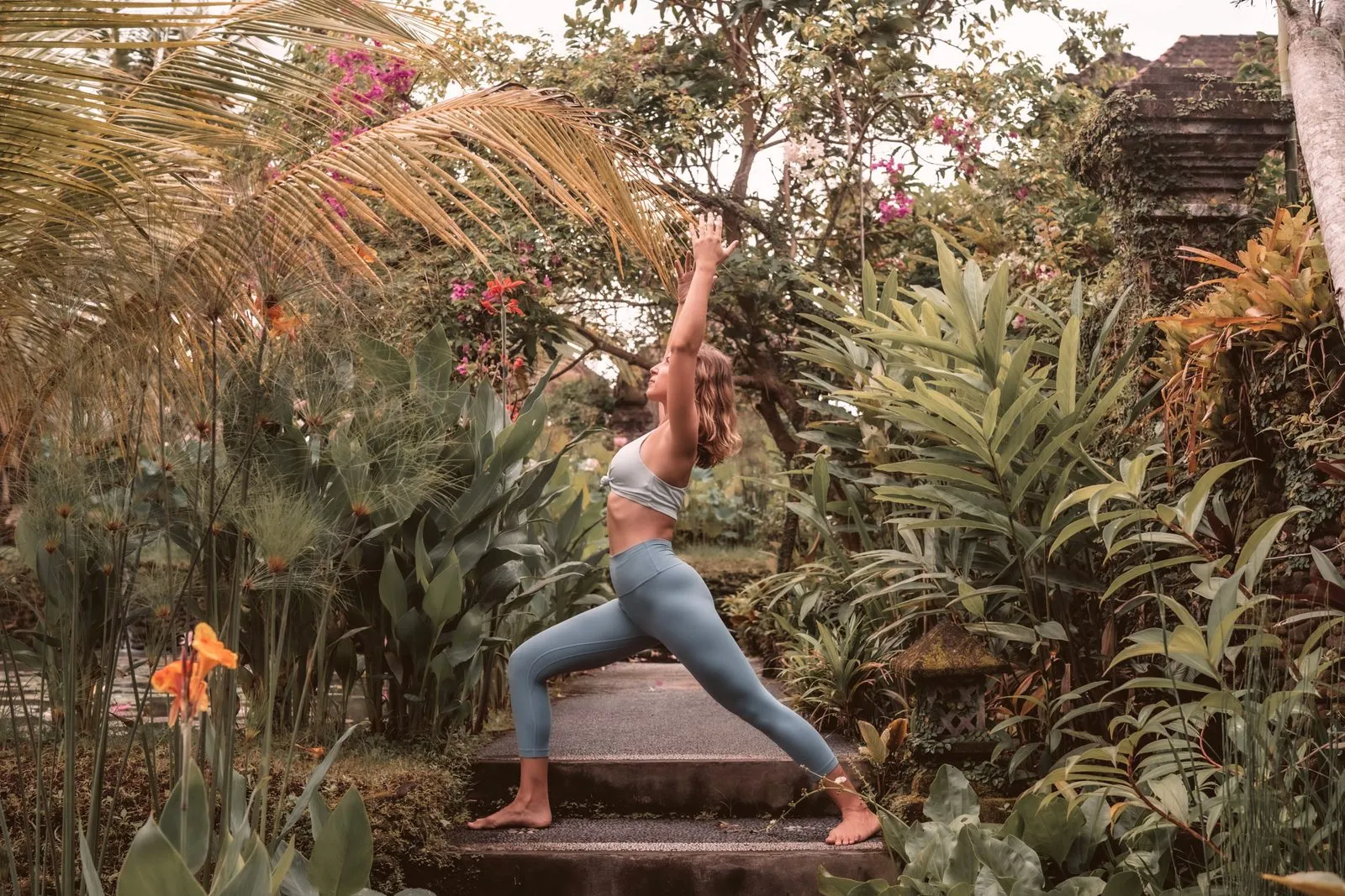 A woman in a high lunge with arms raised on stone steps in a tropical garden, a temple roof behind
