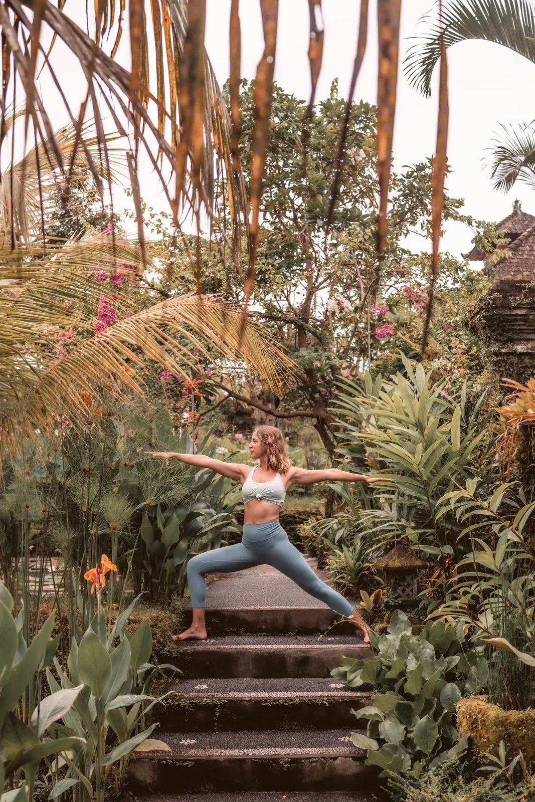 A woman in warrior two on garden steps beneath palm fronds and pink bougainvillea