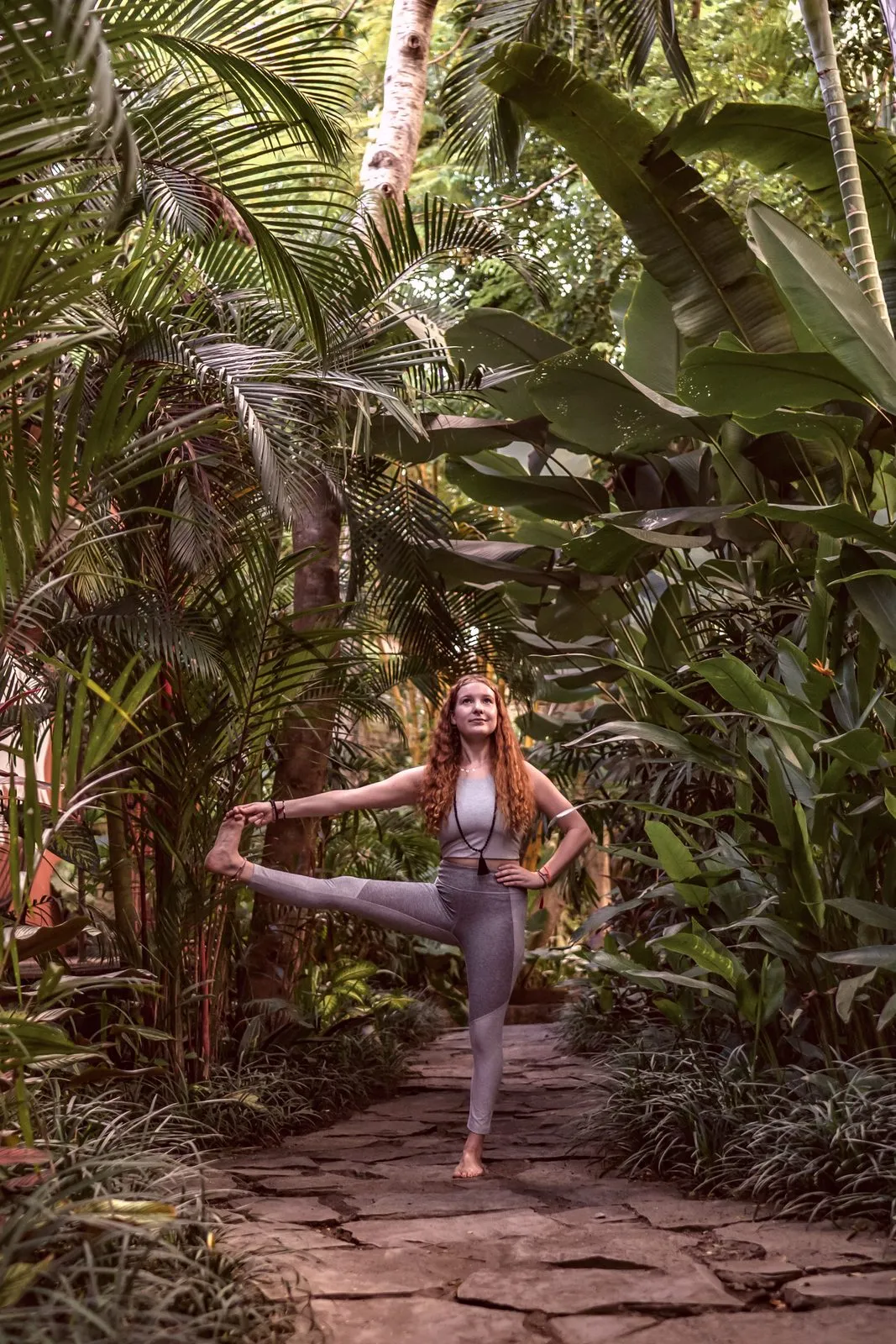 A woman in dancer pose on a stone path through a jungle garden, holding her lifted foot
