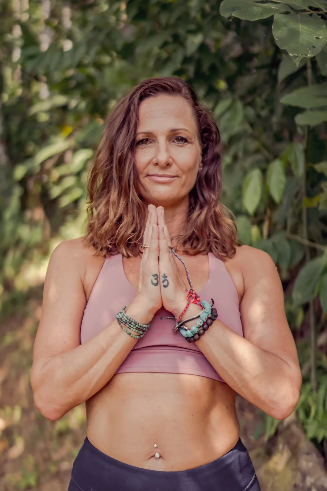 A woman in a pink crop top with her hands in prayer at her heart, palm fronds behind her
