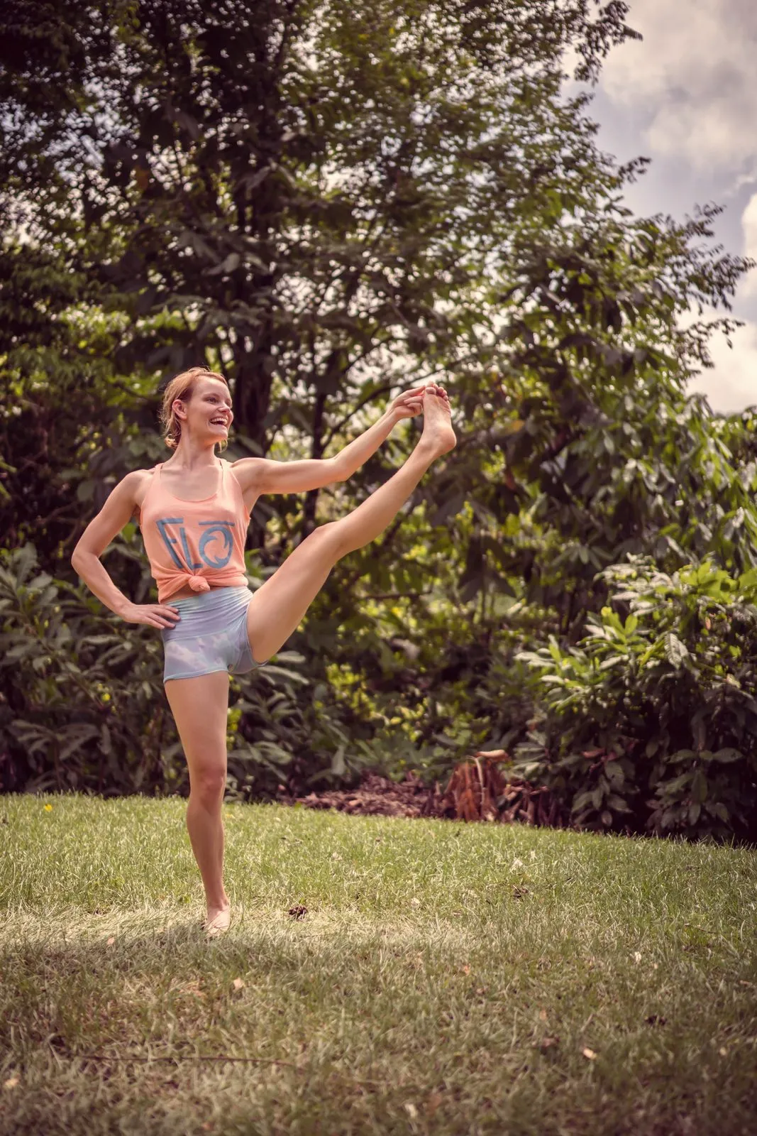 A woman in shorts and a tank top holding her extended leg high in a standing balance on the lawn
