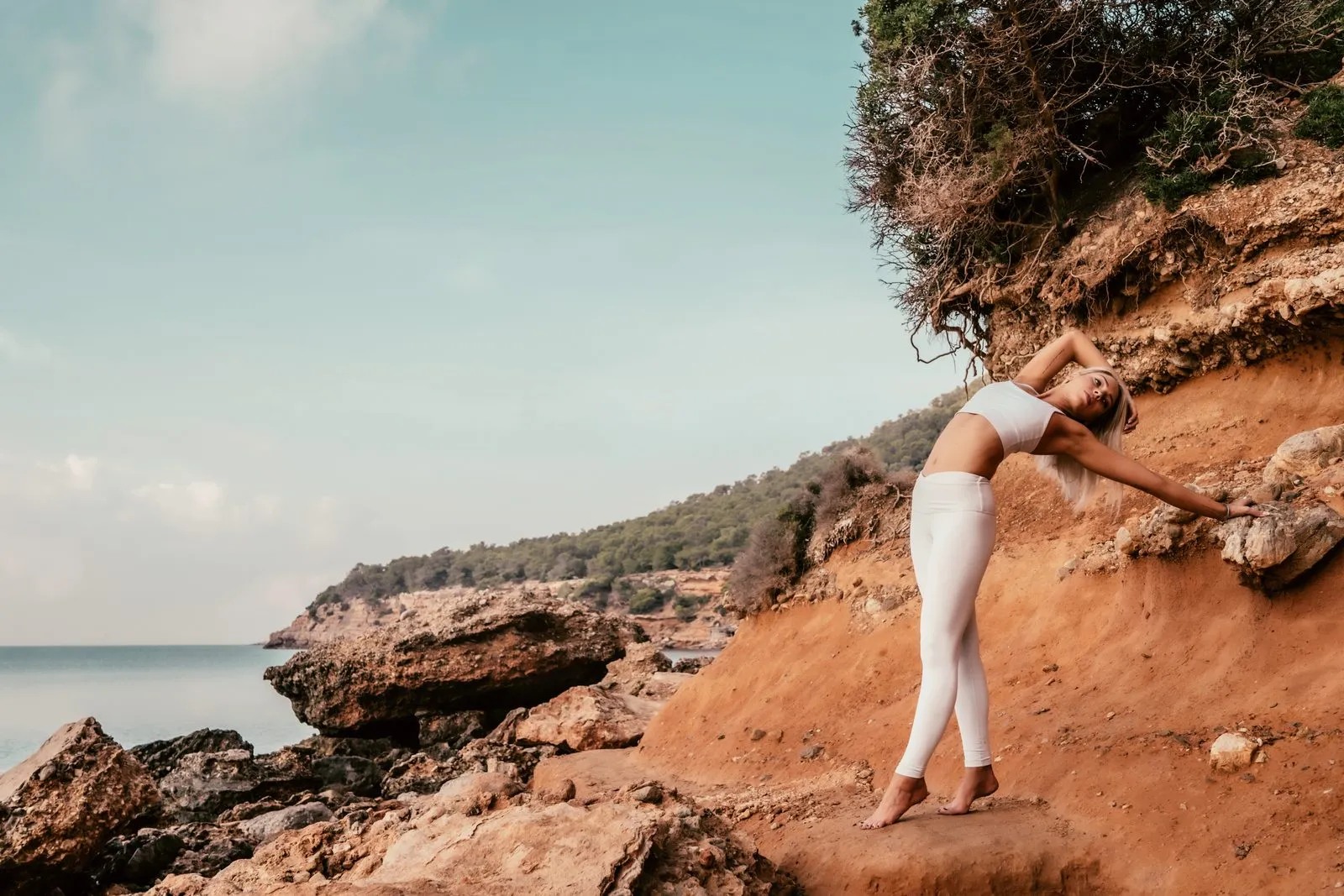 A woman in white doing yoga on red rocks above a calm sea, arms stretched in a lunge