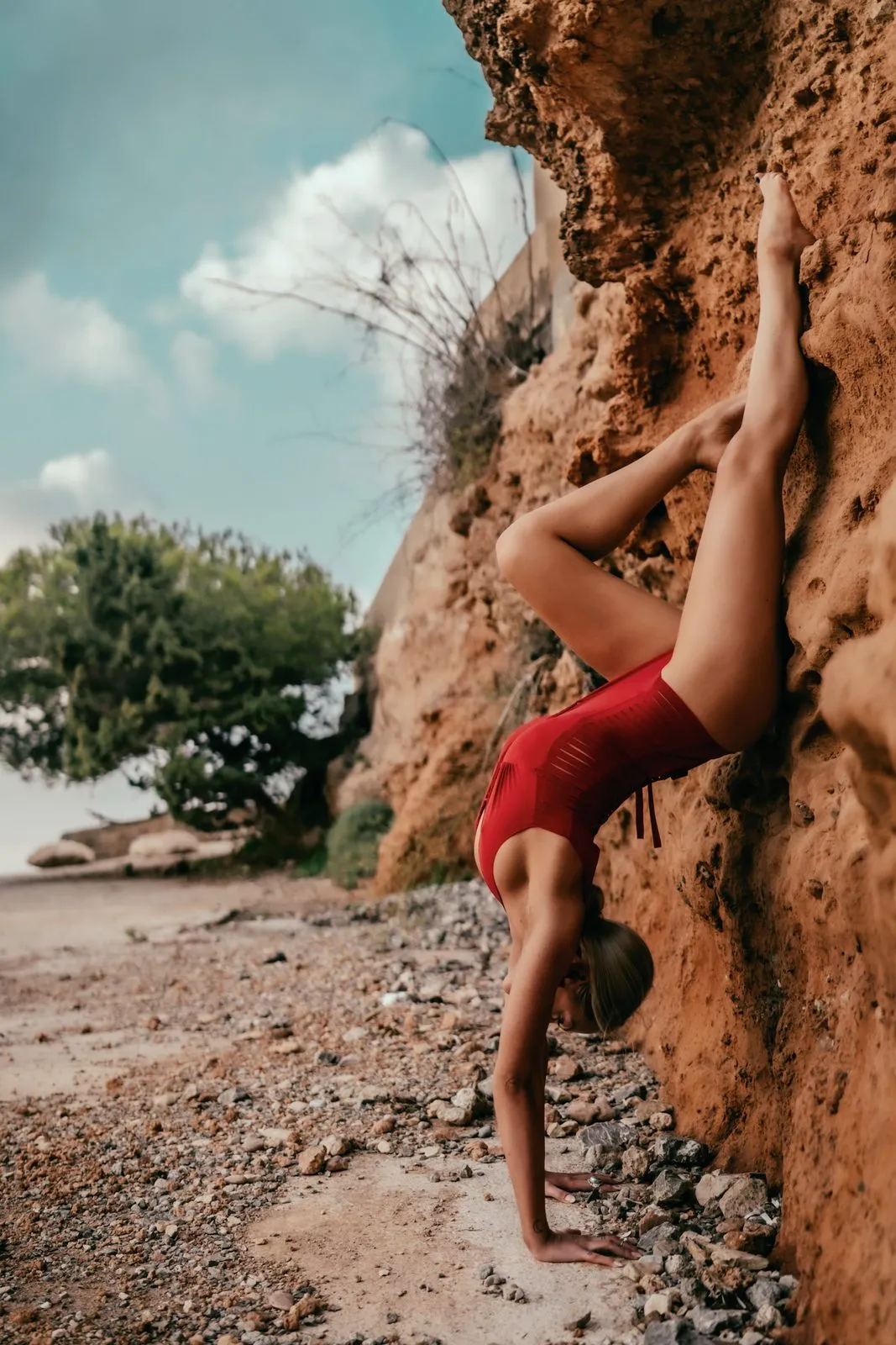 A woman in red in a backbend against a red rock face on a beach, one leg raised