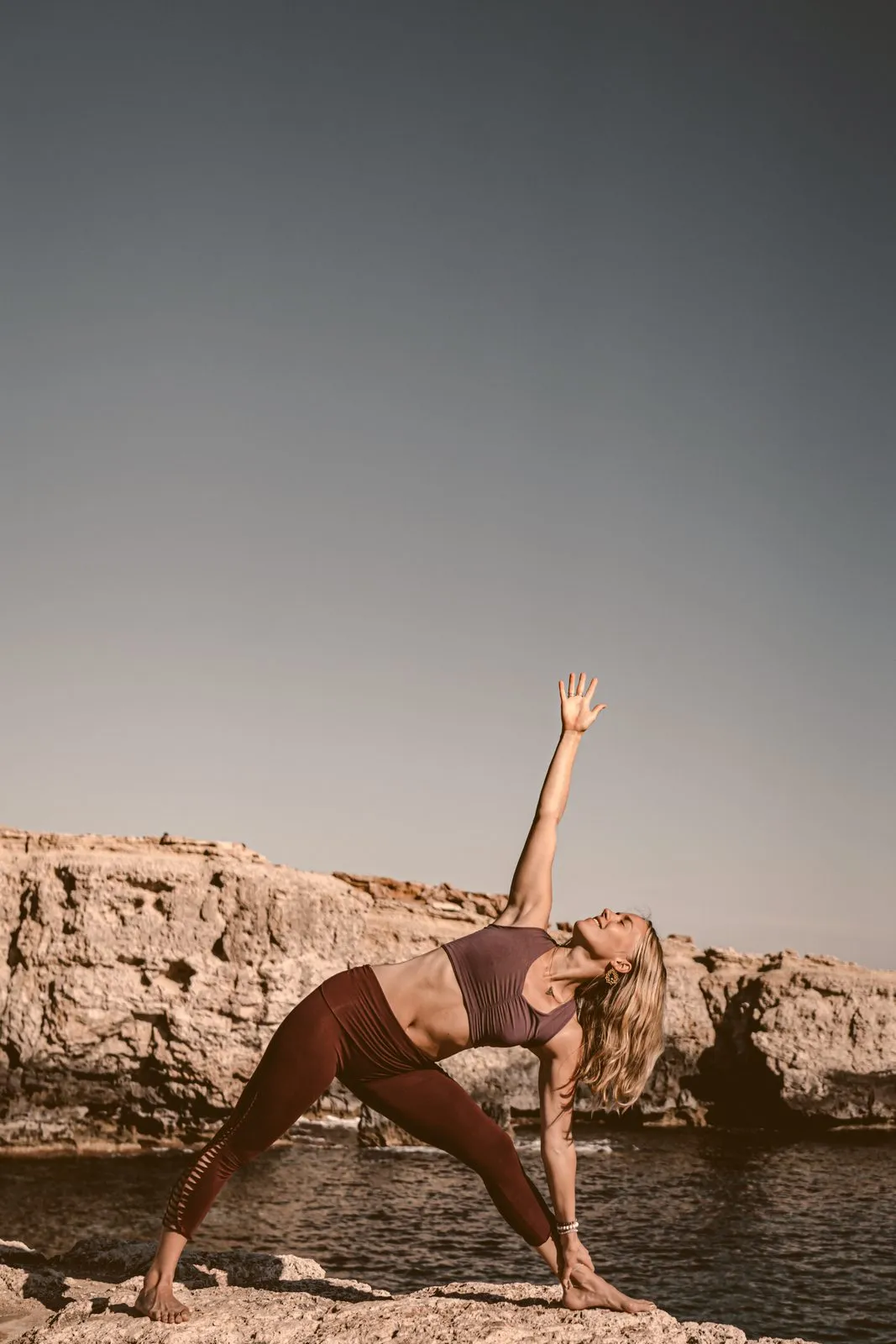 A woman in triangle pose on flat rocks by the sea, one arm pointing to a clear evening sky