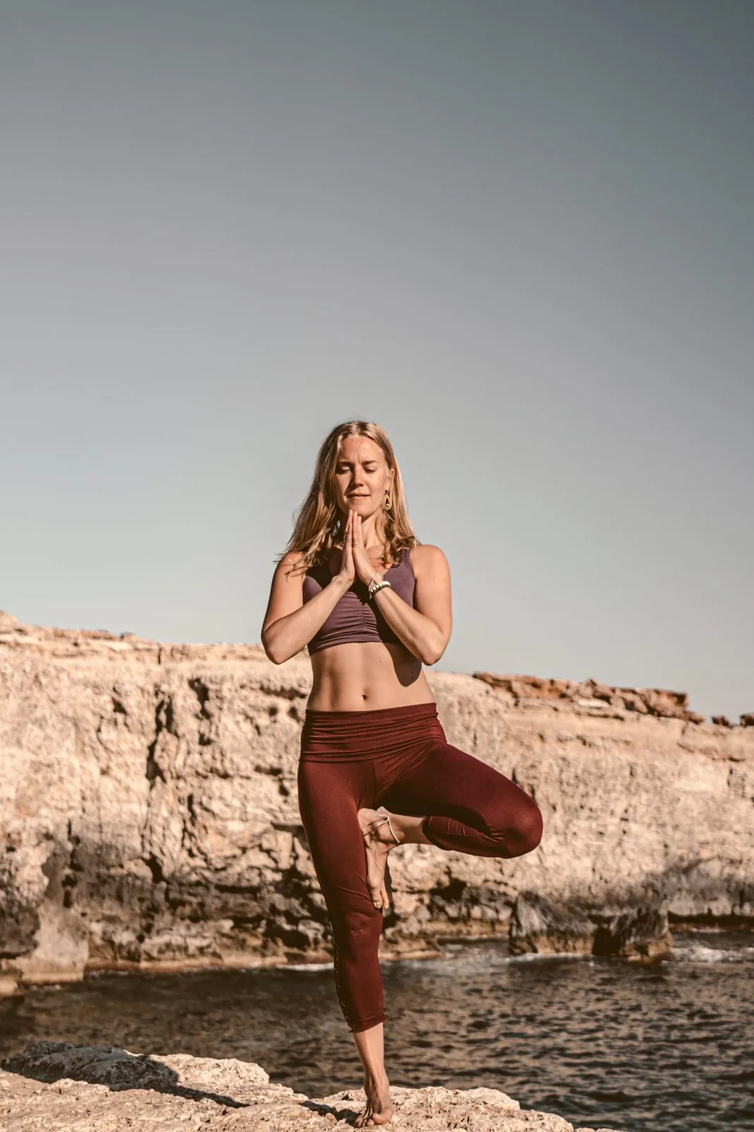 A woman in tree pose with her hands in prayer on a rock shelf, low cliffs and the sea behind her