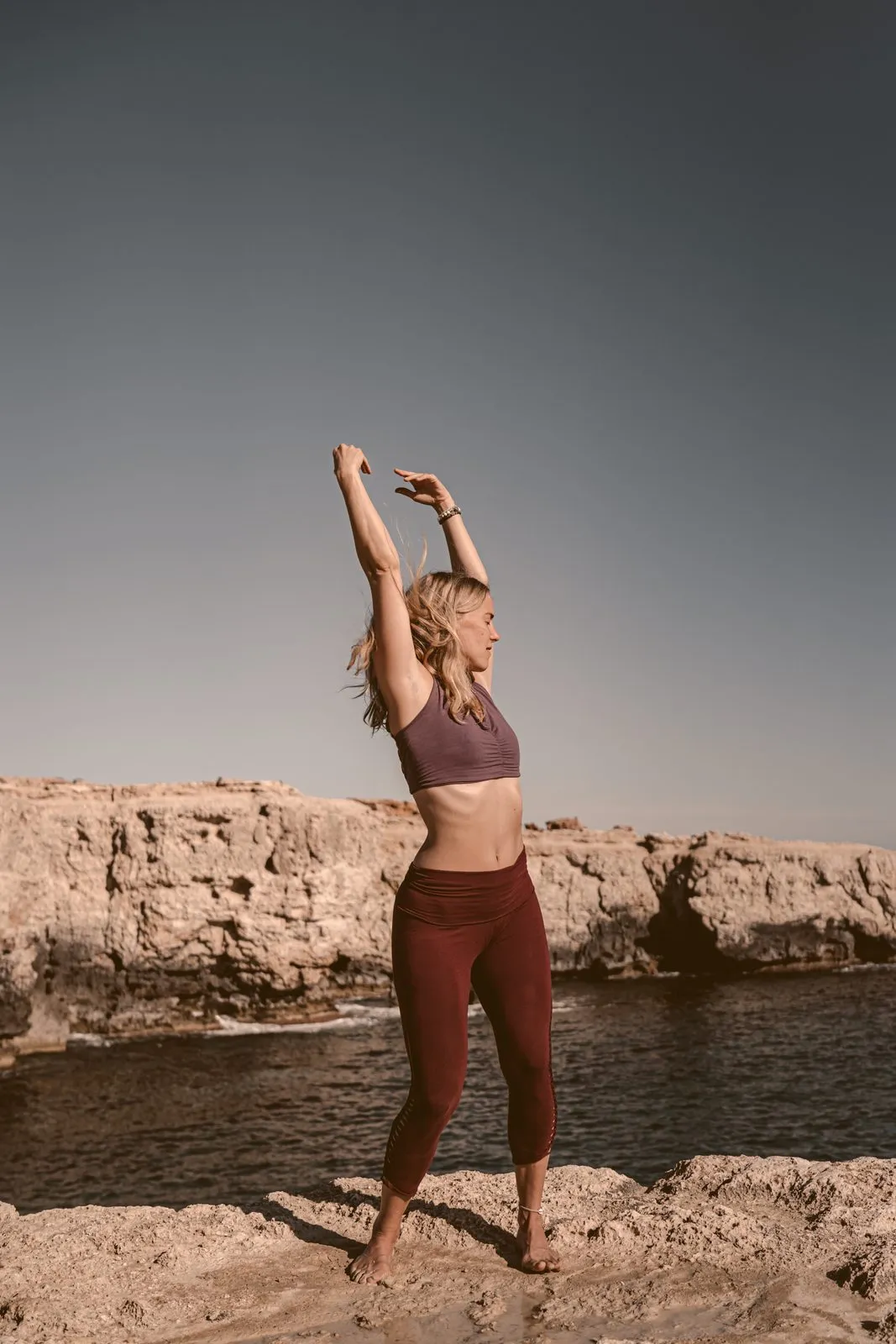 A woman stretching both arms overhead on the rocks at the edge of the sea, in the last of the light