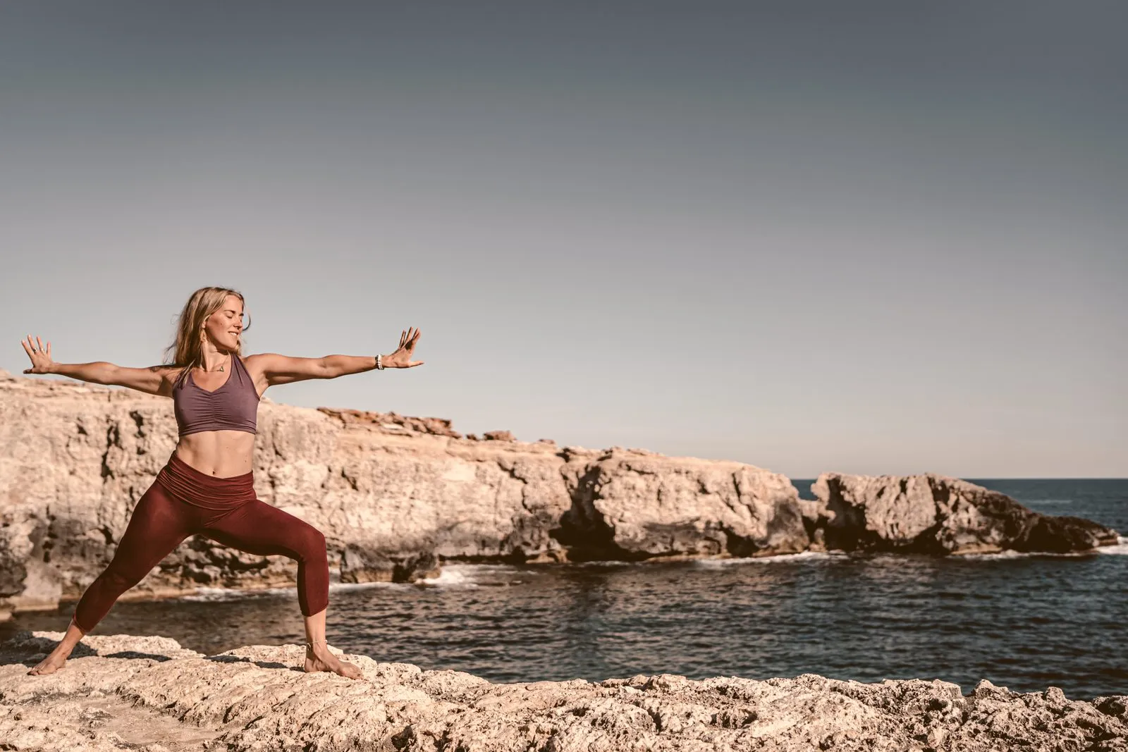 A woman in burgundy leggings and a mauve top in warrior two on a limestone headland, arms wide, the sea behind her